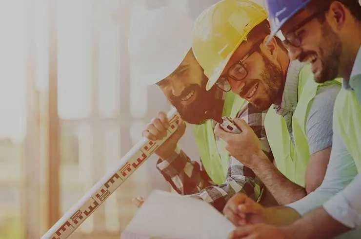 Two smiling construction workers in hard hats review blueprints with a ruler.